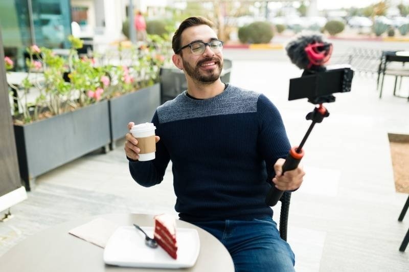 Content creator recording a lifestyle vlog with a smartphone on a tripod while enjoying coffee at an outdoor café.