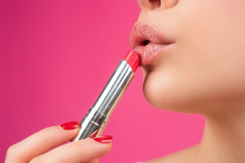 Close-up of a woman applying bright red lipstick against a solid pink background.