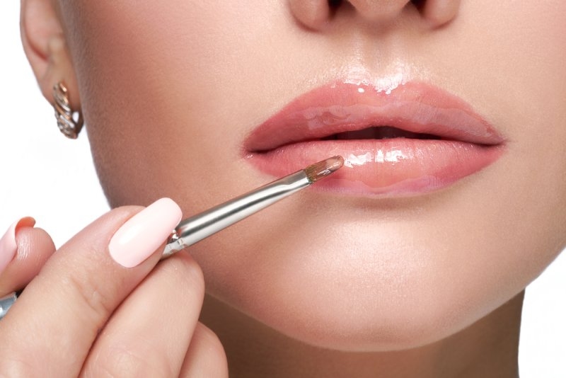 Close-up of a woman applying glossy lip color with a makeup brush, highlighting a shiny, natural-looking lip finish.