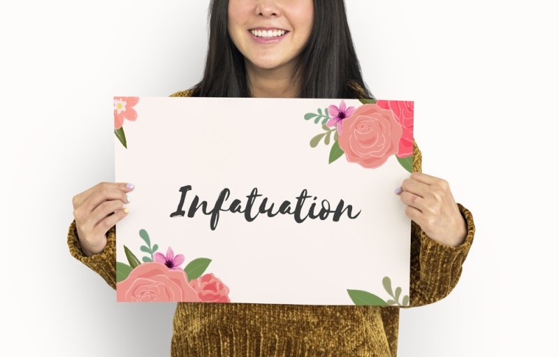 Smiling woman holding a floral sign with the word “Infatuation” against a plain white background.