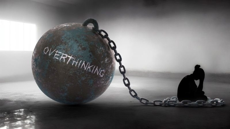 Conceptual illustration of overthinking, showing a person chained to a large metal ball labeled "Overthinking" in a dark, empty space.