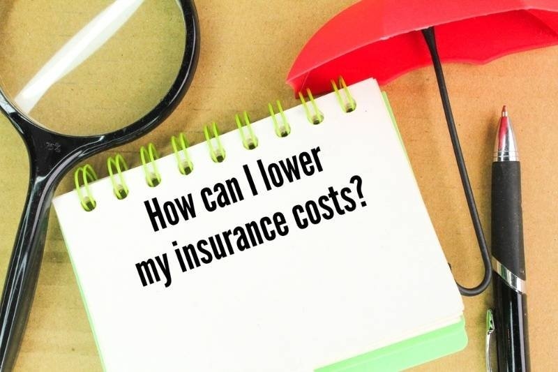 Notepad displaying the question "How can I lower my insurance costs?" surrounded by a magnifying glass, red umbrella, and pen.