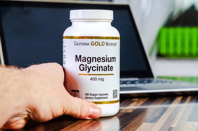 Hand holding a bottle of California Gold Nutrition Magnesium Glycinate 400 mg dietary supplement in front of a laptop.