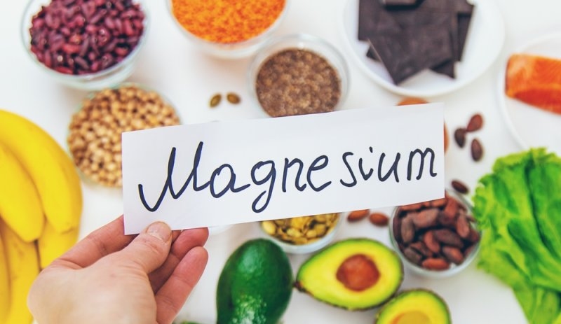Hand holding a card labeled “Magnesium” surrounded by magnesium-rich foods including bananas, avocado, nuts, seeds, beans, and leafy greens.