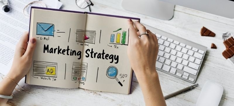 Open notebook featuring a marketing strategy plan with icons for email, website, advertising, SEO, and analytics beside a computer keyboard on a workspace.