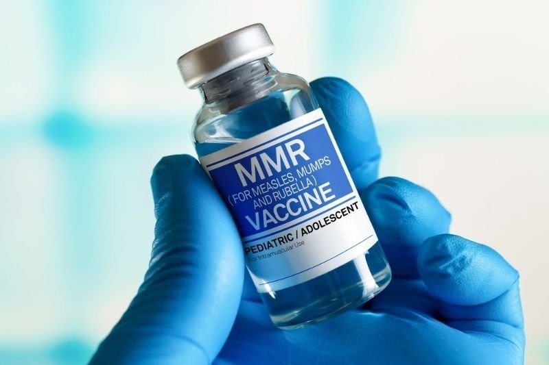 Close-up of a gloved hand holding an MMR vaccine vial labeled for measles, mumps, and rubella immunization.