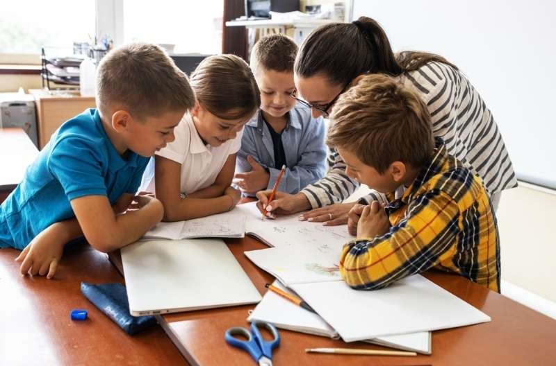 Teacher assisting a group of children working together on school assignments at a table in a classroom.