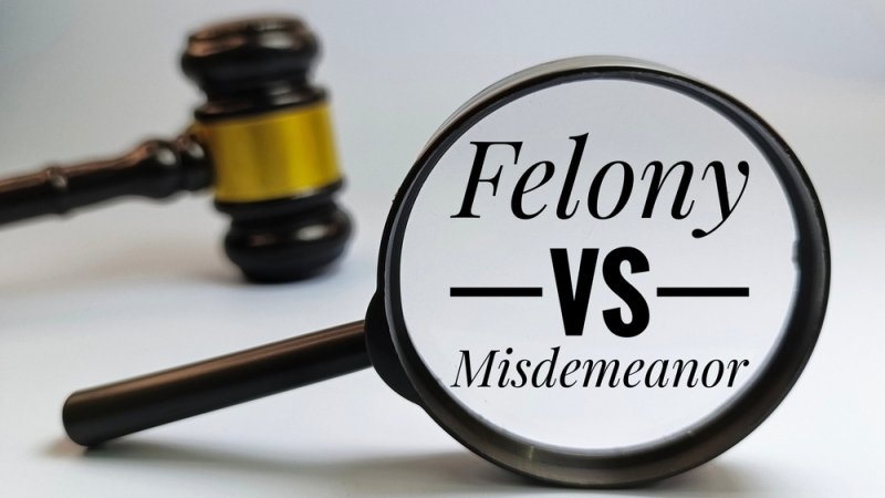 Felony vs Misdemeanor Text Seen Through Magnifying Glass with Blurred Judge Gavel in Background.