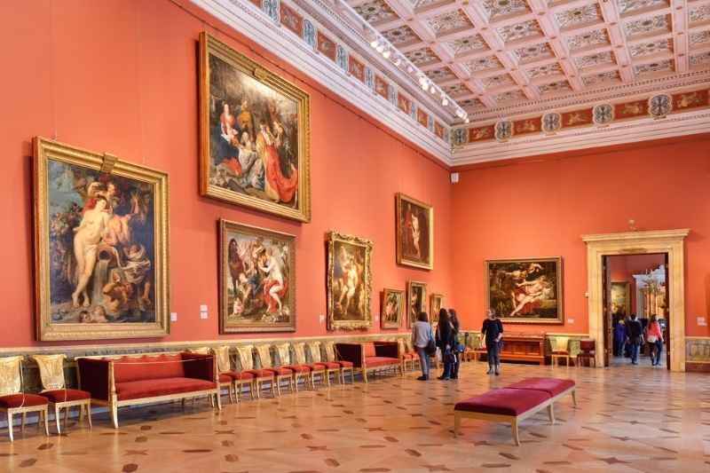 Art gallery interior with large classical paintings on red walls, ornate ceiling, benches, and visitors viewing the artwork.