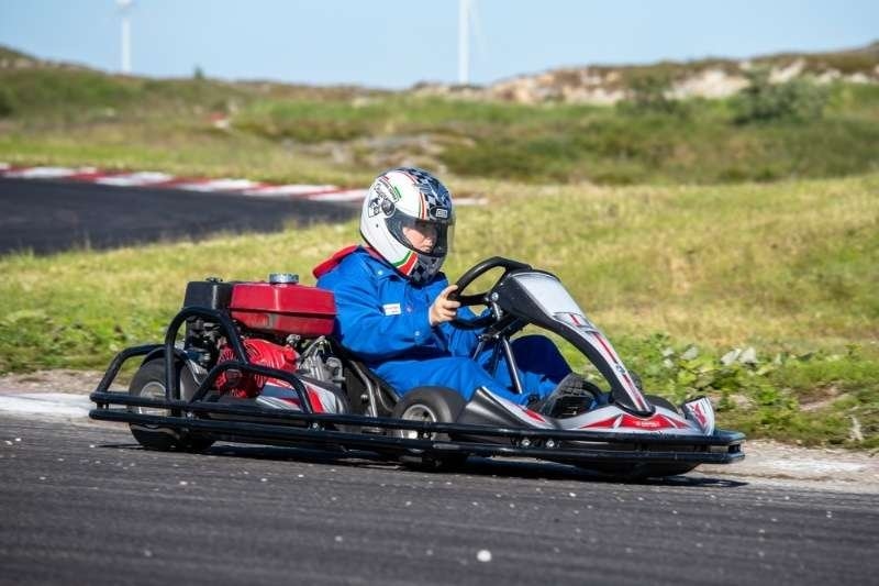 A helmeted driver in a blue racing suit drives a go-kart around an outdoor track, with grassy terrain and wind turbines in the background.