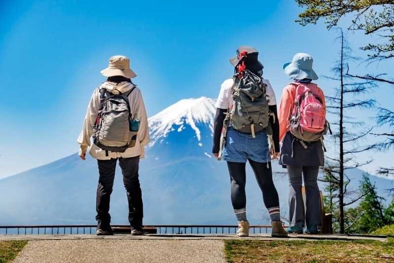 Three hikers with backpacks stand at a viewpoint admiring the snow-covered peak of Mount Fuji on a bright, sunny day.