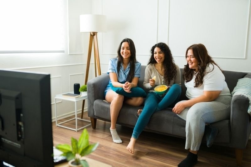 Three friends sitting on a sofa watching TV, smiling and enjoying snacks during a movie night in a cozy living room.