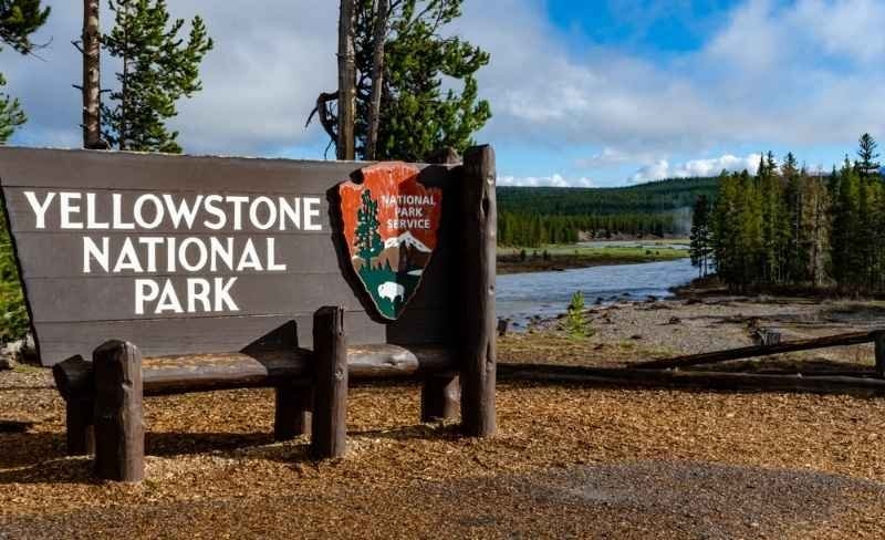 Entrance sign for Yellowstone National Park with a river and evergreen trees in the background.