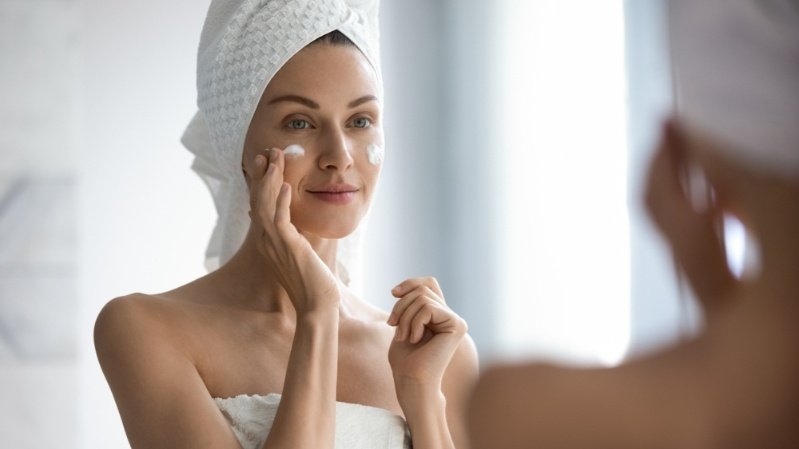Person in a bath towel applying facial moisturizer while looking in a bathroom mirror during a skincare routine.