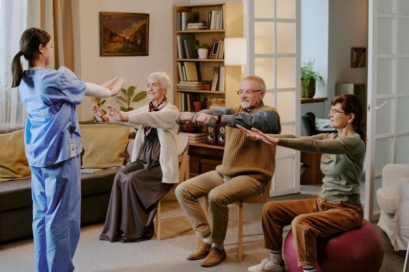 Healthcare professional guiding a small group of seniors performing seated stretching exercises in a living room setting.