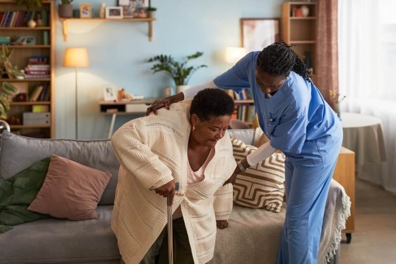 Healthcare worker helping an older woman with a walking cane stand up from a sofa at home.