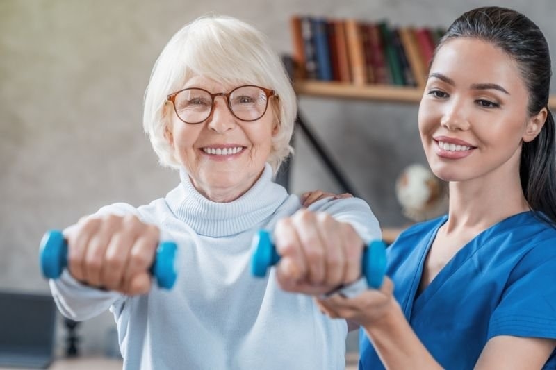 Older woman exercising with small dumbbells while a healthcare professional assists her.