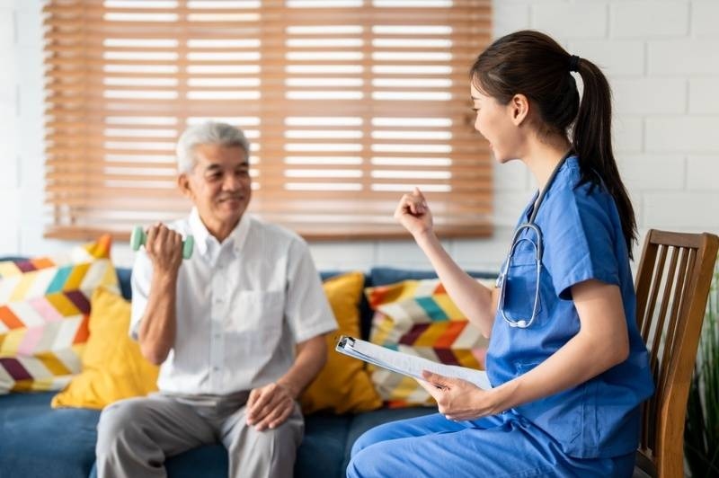 Nurse assisting an elderly man with a light dumbbell exercise during a home visit, holding a clipboard.