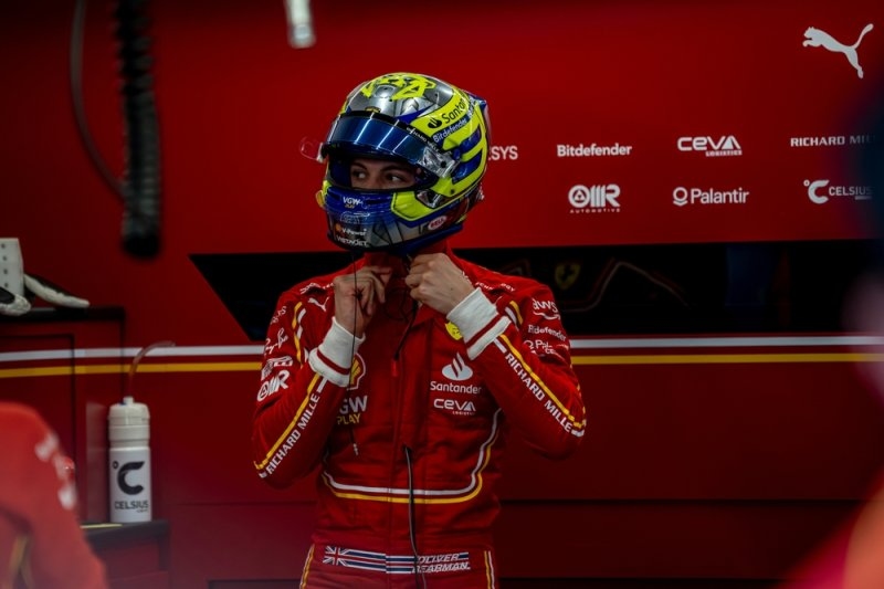 Formula 1 driver wearing a red racing suit and helmet inside a Ferrari garage.