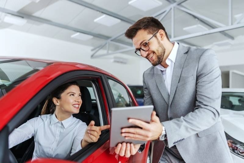 Insurance or sales representative showing insurance information on a tablet to a customer seated inside a red car at a dealership.