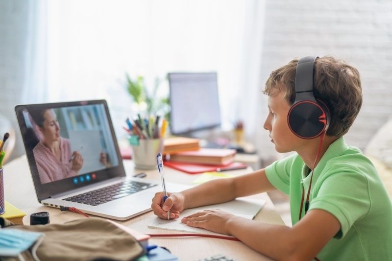 Boy wearing headphones while attending an online class and taking notes at a desk.