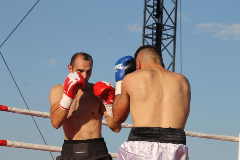 Two male boxers wearing red and blue gloves face each other in an outdoor boxing ring under a clear blue sky.