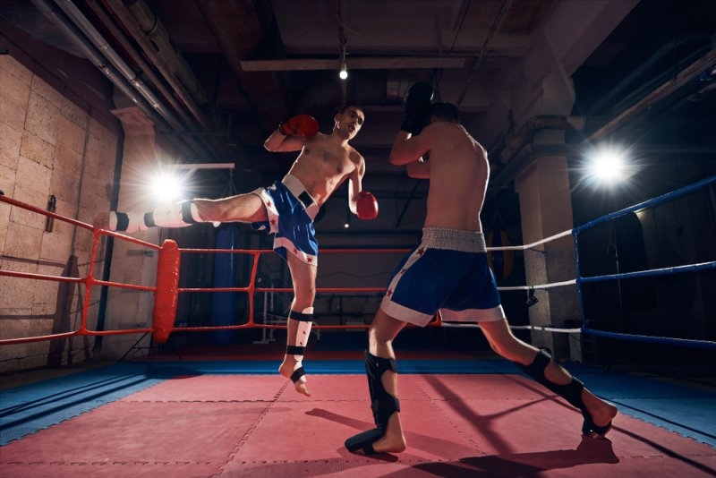 Two male fighters wearing boxing gloves and protective gear sparring inside an indoor boxing ring under bright overhead lights.