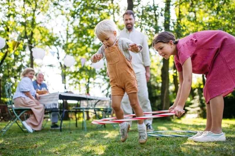 Young child playing with a hula hoop outdoors while family members watch and participate in a backyard gathering.