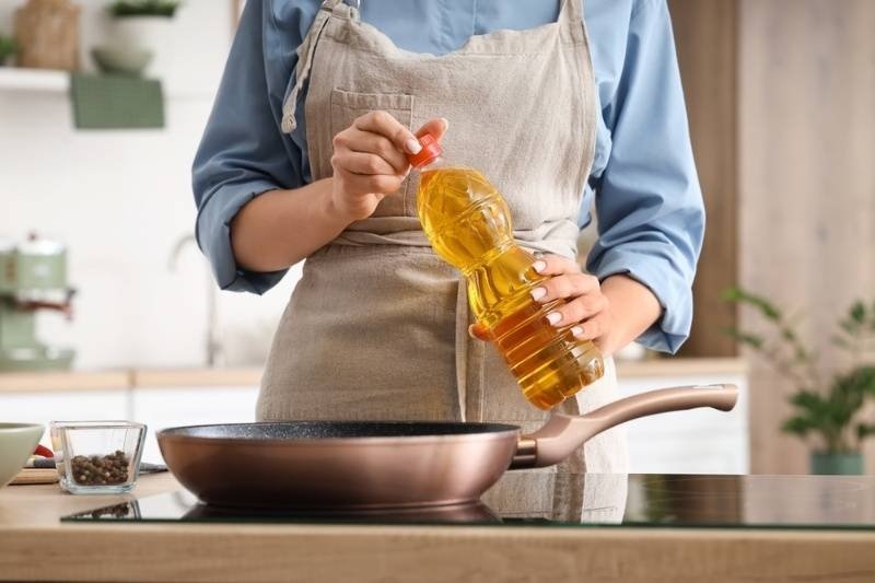 Person holding a bottle of cooking oil above a frying pan in a kitchen.