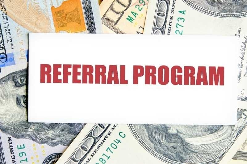 A white sign reading “REFERRAL PROGRAM” is placed over a background of U.S. dollar bills.