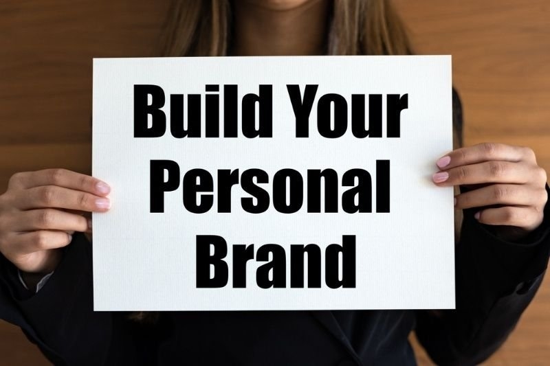 A person in business attire holds a white sign that reads “Build Your Personal Brand.”