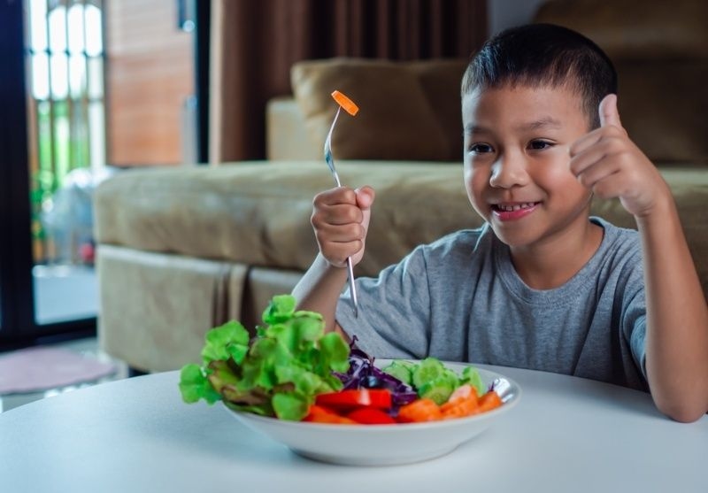 Smiling child giving a thumbs up while holding a fork and eating a bowl of fresh salad.