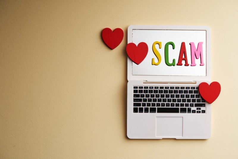 White laptop displaying the word “SCAM” in colorful letters, surrounded by red heart decorations on a beige background.