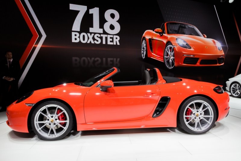 Orange Porsche 718 Boxster convertible displayed at an auto show, with silver alloy wheels, red brake calipers, and a large “718 Boxster” promotional backdrop.