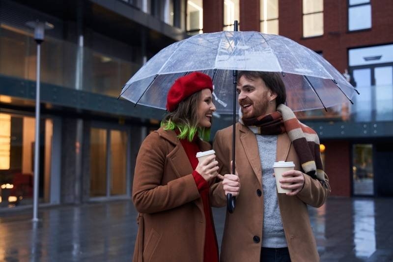 Smiling couple walking under a transparent umbrella while holding takeaway coffee cups on a rainy day.