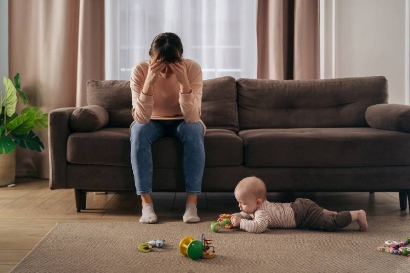 A distressed mother sitting on a sofa with her head in her hands while her baby plays on the floor.