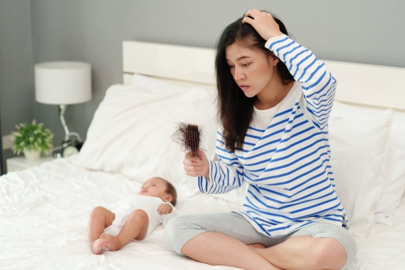 New mother inspecting hair loss on a hairbrush while sitting beside her baby.