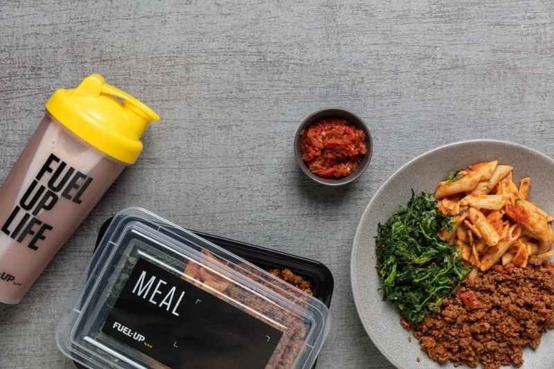 Meal prep containers with a protein shake, lean meat, pasta, and leafy greens.