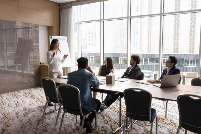 A presenter speaking to a team seated around a conference table in a modern office meeting room.