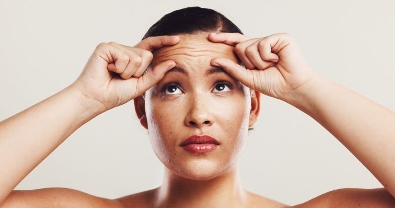 Woman gently pulling the skin on her forehead while looking upward, illustrating wrinkles, aging concerns, or anti-aging skincare.