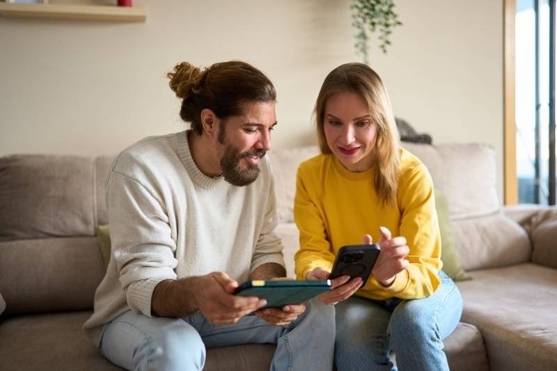 A man and woman sitting together on a couch and looking at a smartphone and tablet.