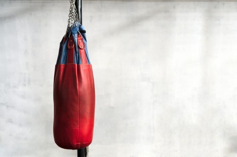 Red and blue heavy punching bag hanging in an empty gym with concrete wall background.