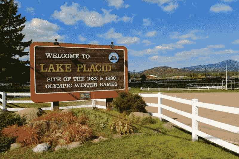 Welcome sign for Lake Placid, New York, highlighting the 1932 and 1980 Olympic Winter Games, with mountains and open fields in the background.