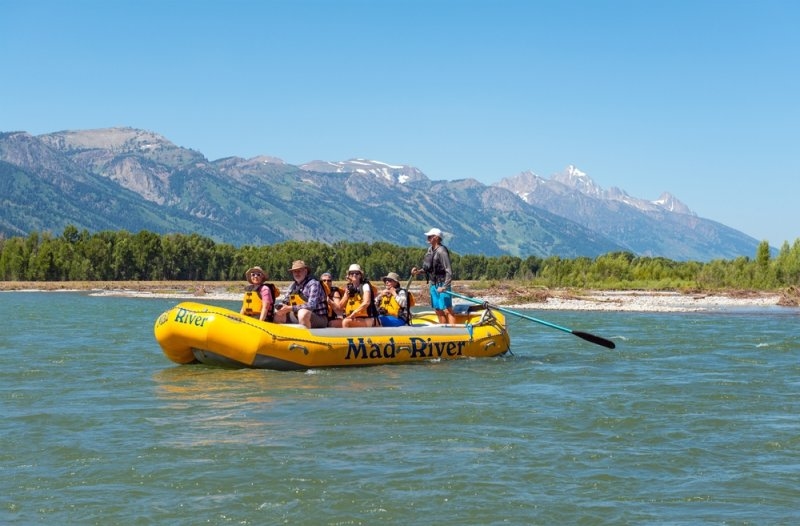 Group of people wearing life jackets riding a yellow inflatable raft on a river with mountains in the background.