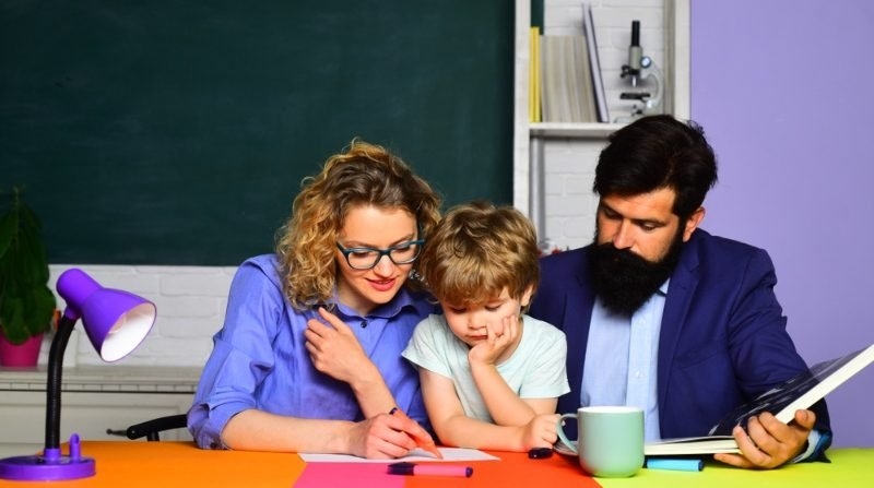 Parents helping a young child with schoolwork at a desk.