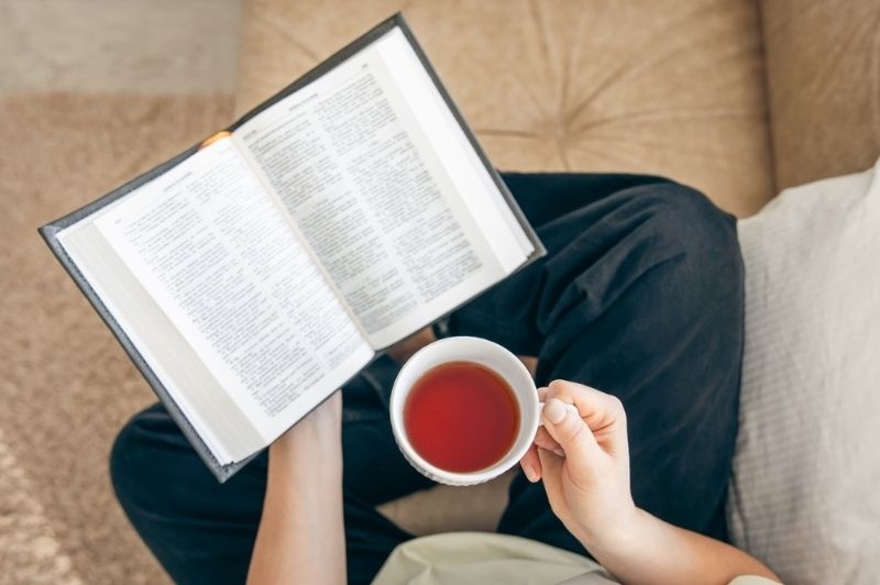 Person sitting comfortably while reading a book and holding a cup of tea, representing a relaxing reading moment.