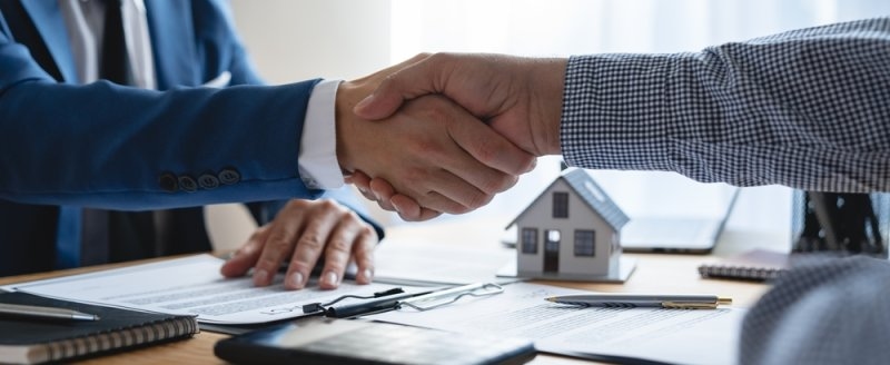 Two people shaking hands over real estate documents with a small house model on the desk.