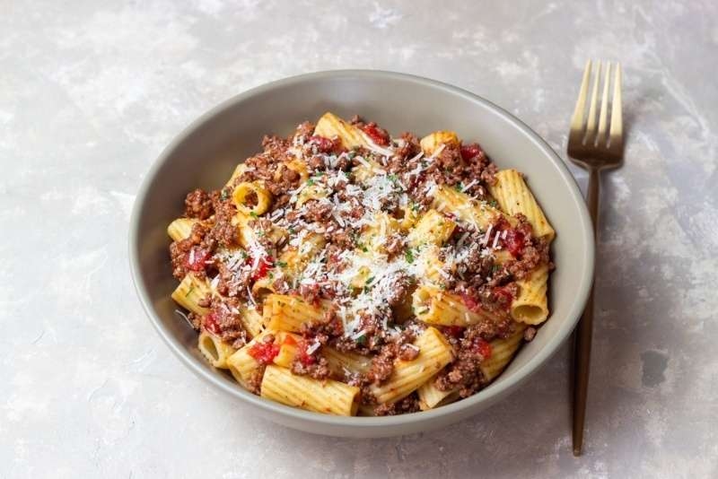 Plate of rigatoni pasta topped with meat sauce and grated parmesan cheese beside a fork on a gray background.