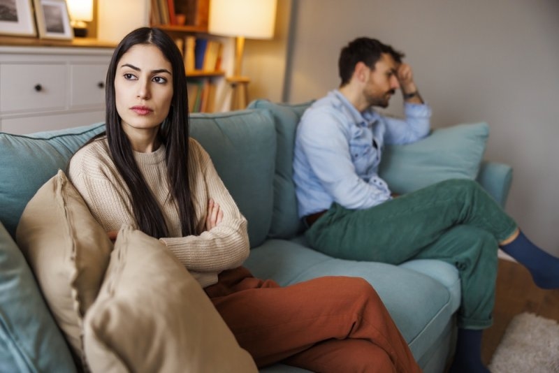 Couple sitting apart on a sofa, with the woman looking away with crossed arms and the man appearing upset in the background.