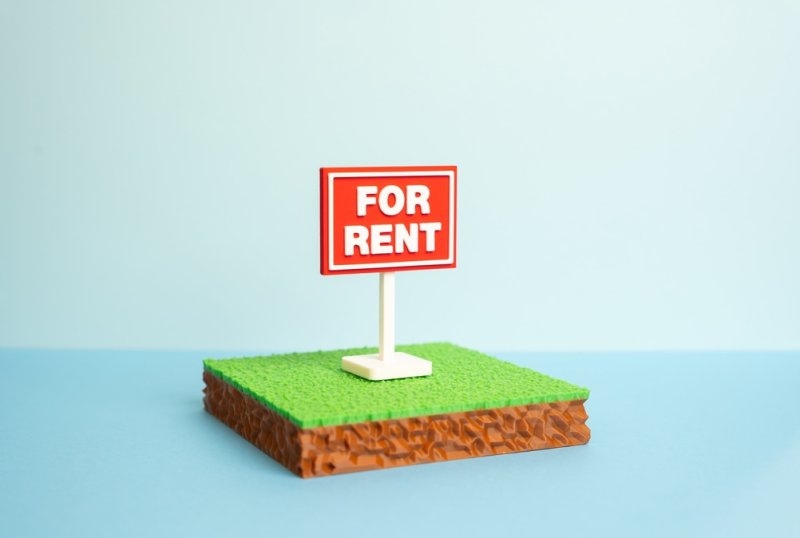 A miniature “For Rent” sign stands on a small patch of grass, representing rental property or a rental business.